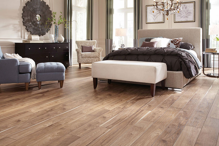 Large open bedroom decorated with a bed on a modern frame, a ceiling mounted pendant chandelier, a large dark stained wood dresser, two sitting chairs, and two ottomans on light colored engineered wood floors.