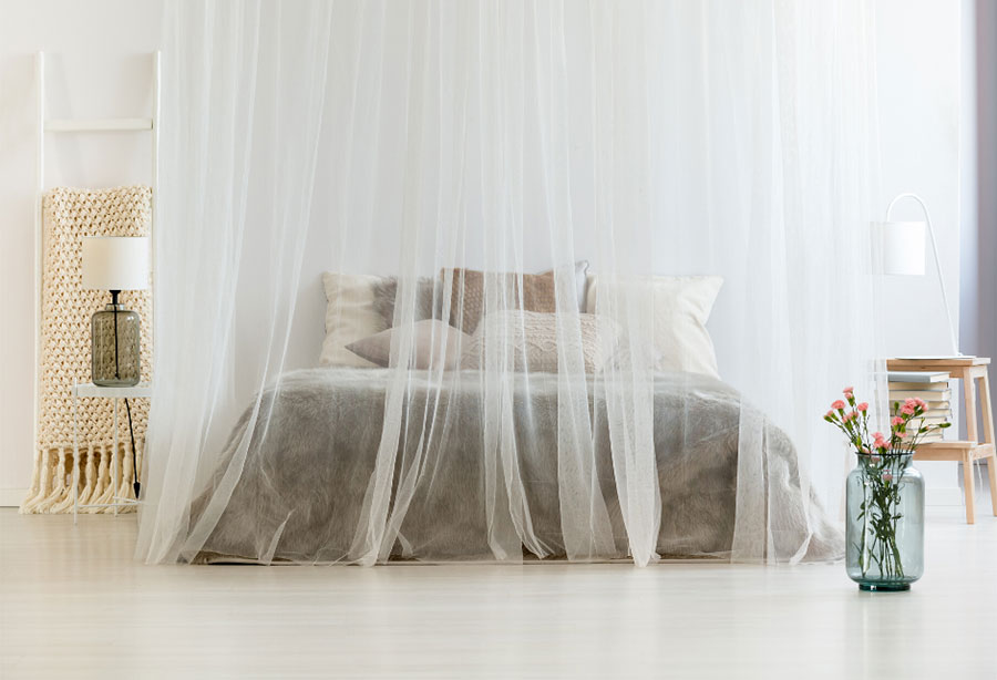 Sheek white bedroom furnished with a canopy bed draped in mosquito netting and a vase with pink flowers.